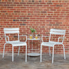 Two white Luxembourg armchairs, placed on either side of a round side table, against a brick wall background.