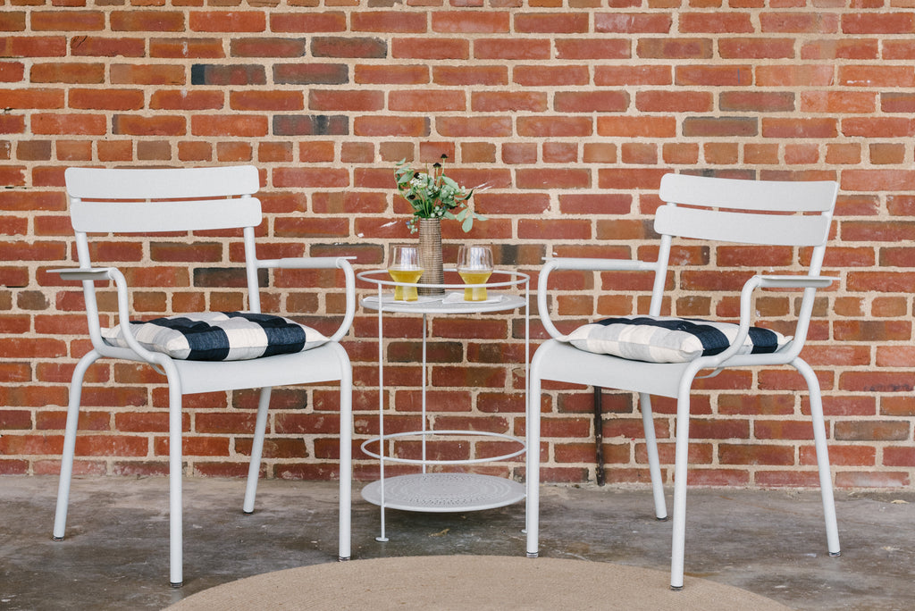 Two white Luxembourg armchairs with cushions, placed on either side of a round gside table, against a brick wall background.