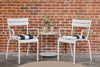 Two white Luxembourg armchairs with cushions, placed on either side of a round gside table, against a brick wall background.