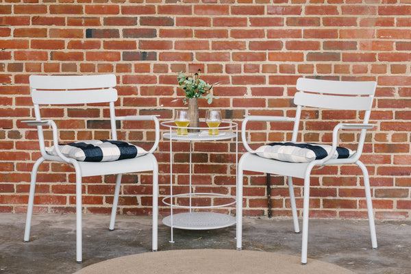 Two white Luxembourg armchairs with cushions, placed on either side of a round gside table, against a brick wall background.