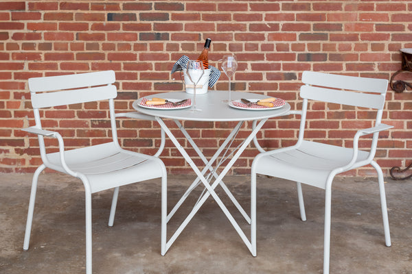 Two white Luxembourg armchairs, placed on either side of a round table, against a brick wall background.