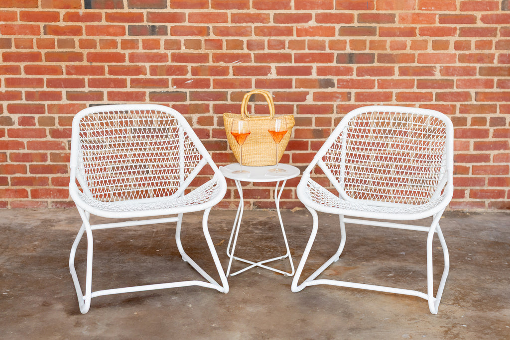 Two white low armchairs with a criss-cross weave on an aluminum frame, accompanied by a small matching round table, set against a brick wall on a concrete surface.