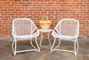 Two white low armchairs with a criss-cross weave on an aluminum frame, accompanied by a small matching round table, set against a brick wall on a concrete surface.