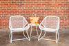 A white metal side table with a geometric design, accompanied by two white chairs and a wicker tote bag with a glass of wine and beer on the table.