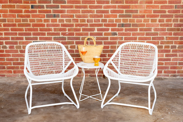 A white metal side table with a geometric design, accompanied by two white chairs and a wicker tote bag with a glass of wine and beer on the table.