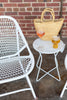 A white metal side table with a geometric design, accompanied by a chair and a wicker tote bag with a glass of wine and beer on the table.