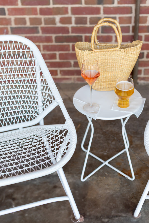 One white low armchair with a criss-cross weave on an aluminum frame, accompanied by a small matching round table, set against a brick wall on a concrete surface.