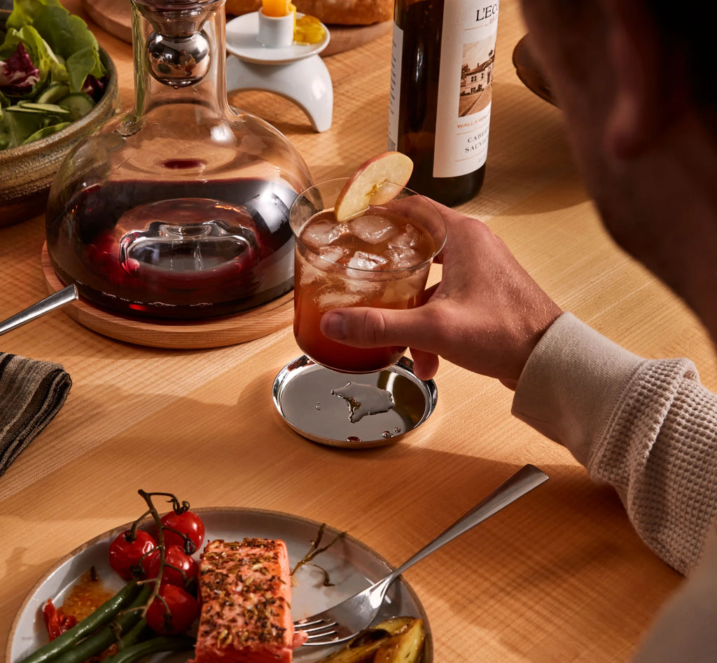 One polished stainless steel coaster with a shallow lip and central anti-slip pad, displayed on a wooden table.