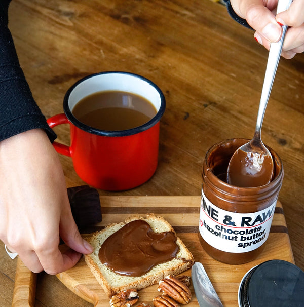Person spreading chocolate spread on toast with a jar of 'FINE & RAW' chocolate hazelnut butter in the background.