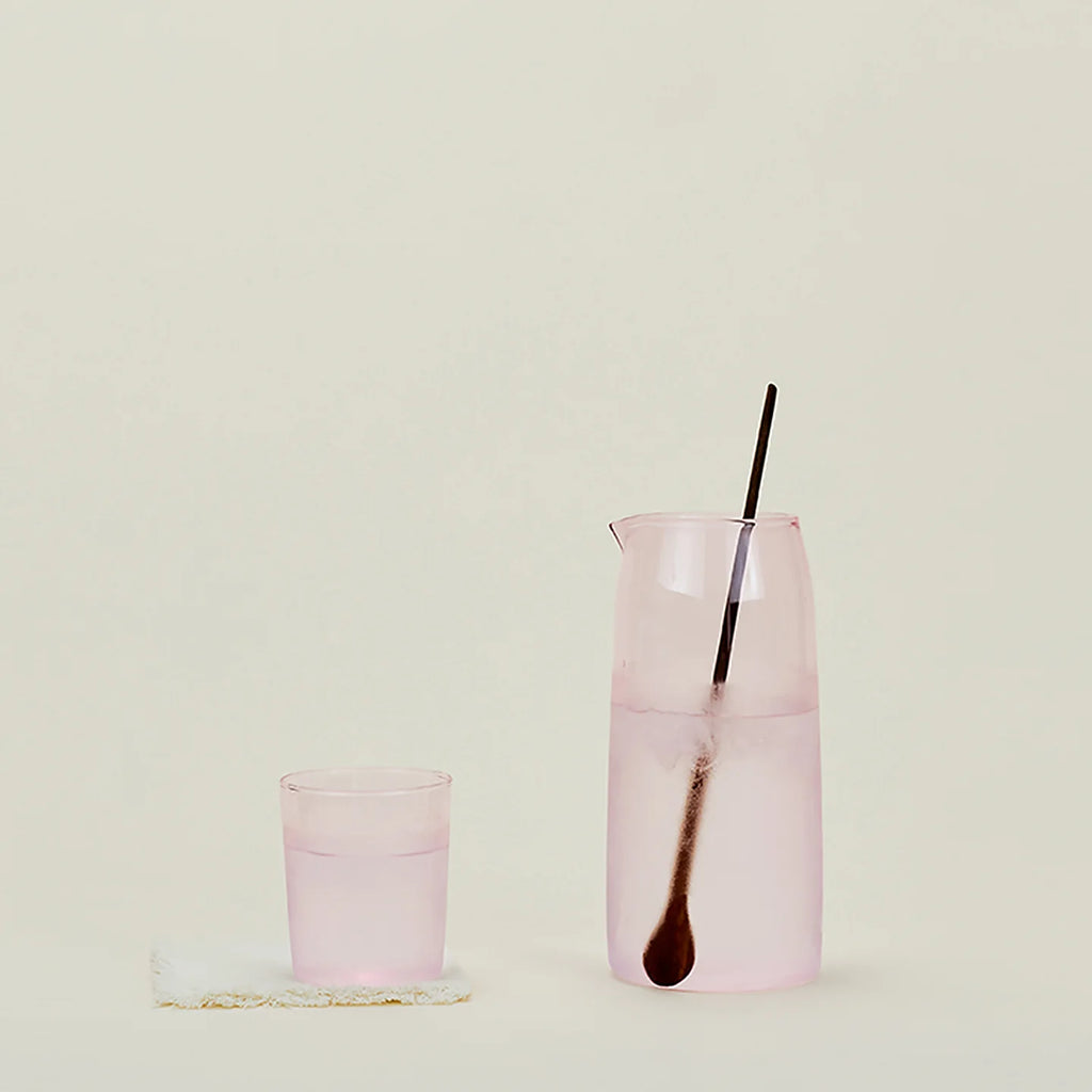 A pink, glass pitcher with a walnut spoon-stirrer, placed next to a small, blush cup on a neutral background.