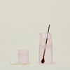 A pink, glass pitcher with a walnut spoon-stirrer, placed next to a small, blush cup on a neutral background.
