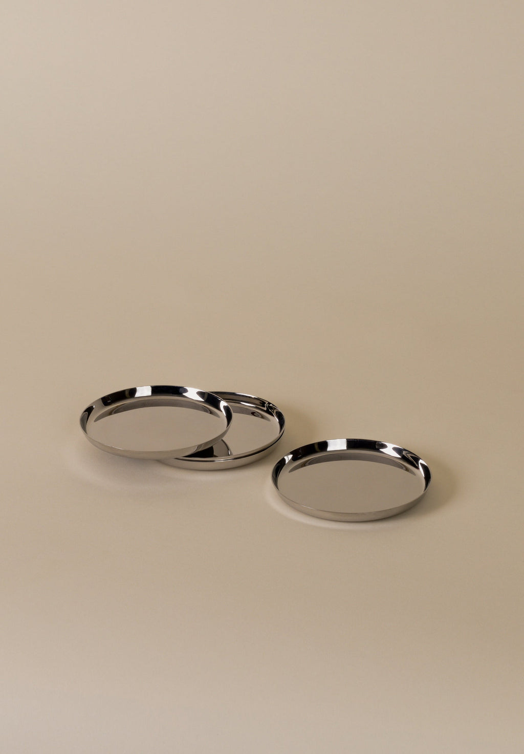 Three polished stainless steel coasters with a shallow lip and central anti-slip pad, displayed on a neutral background.