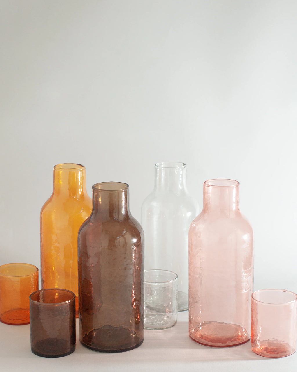 A collection of handblown hammered glassware including various colored glass bottles and tumblers with a textured, pebbled surface.