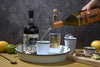 Person pouring a bottle of liquor into a glass with tonic water on a table and a serving tray.
