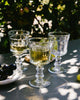 Three clear glass goblets with a drink on a reflective surface with greenery in the background