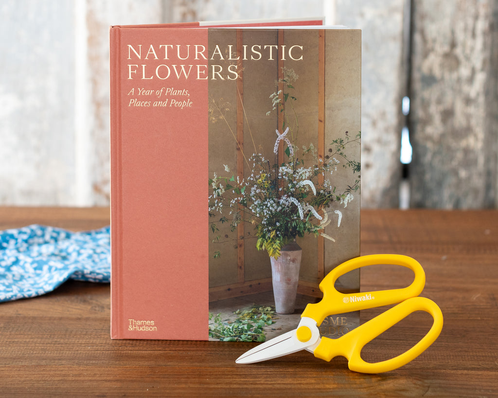 The book 'Naturalistic Flowers: A Year of Plants, Places and People' by Thames & Hudson, displayed on a wooden table next to a pair of yellow-handled scissors.