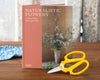 The book 'Naturalistic Flowers: A Year of Plants, Places and People' by Thames & Hudson, displayed on a wooden table next to a pair of yellow-handled scissors.