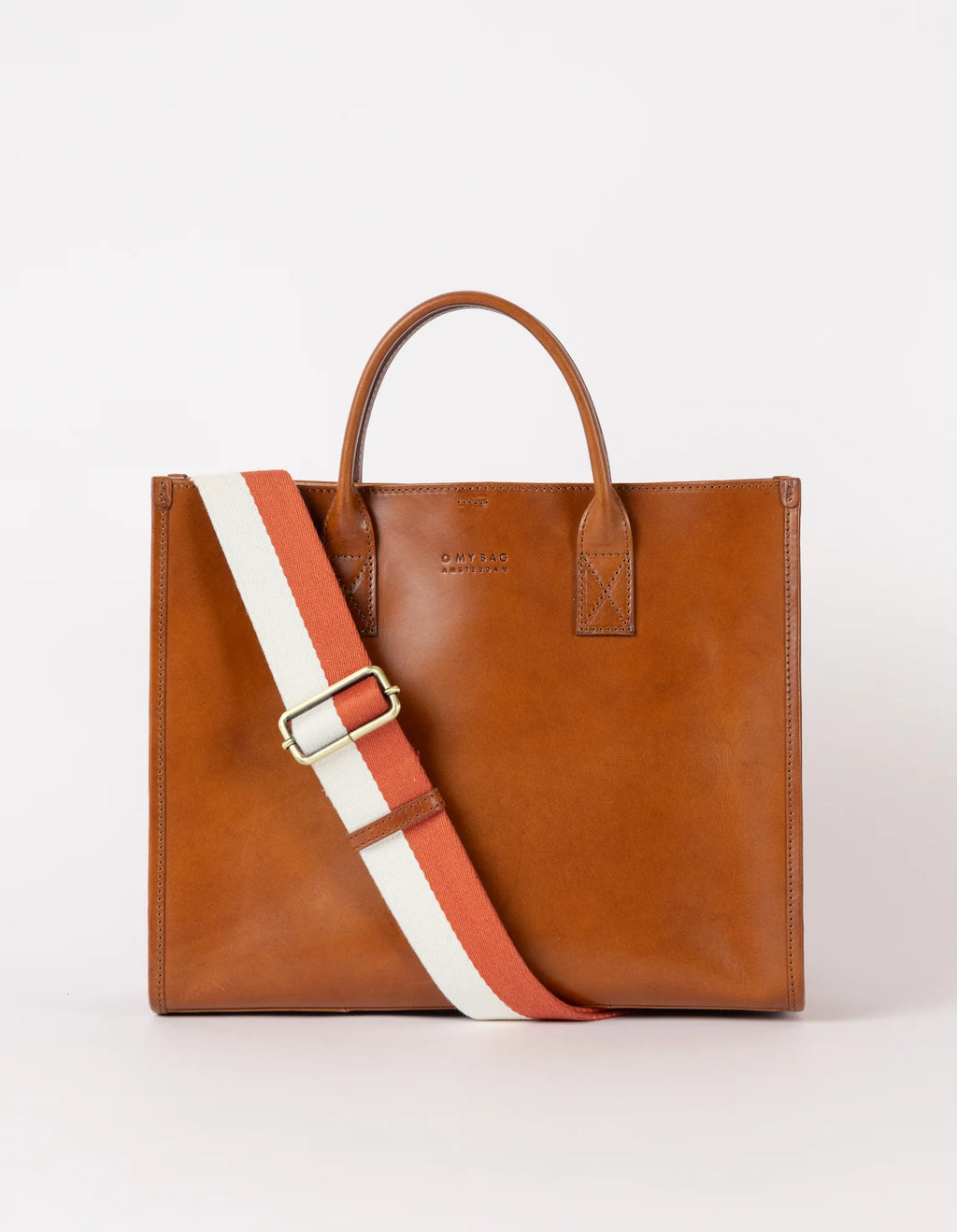 A brown leather tote bag with a structured silhouette, featuring a white and red adjustable strap, two handles, and a minimalist latch closure.