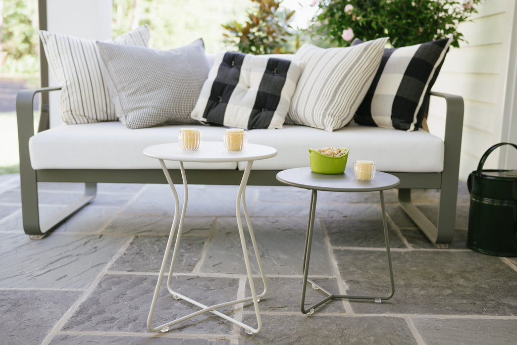 A modern, gray Cocotte Stool with a steel rod frame and a steel sheet seat placed on a stone tiled floor next to a white sofa with striped cushions and a small round table.