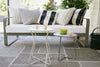 A modern, gray Cocotte Stool with a steel rod frame and a steel sheet seat placed on a stone tiled floor next to a white sofa with striped cushions and a small round table.
