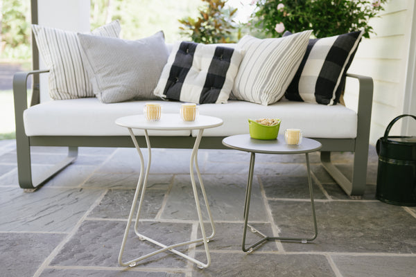 A modern, gray Cocotte Stool with a steel rod frame and a steel sheet seat placed on a stone tiled floor next to a white sofa with striped cushions and a small round table.