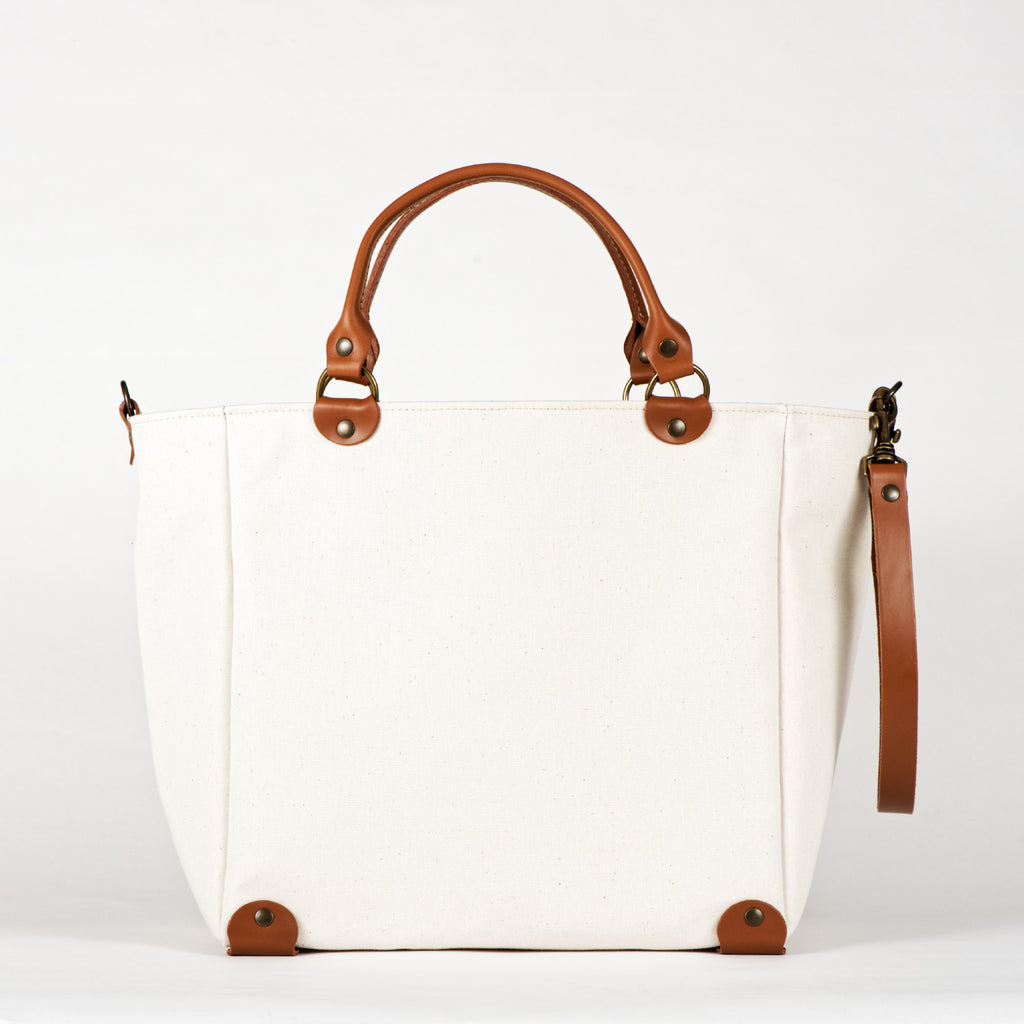 A blanc cotton tote bag with brown leather handles and a removable brown leather strap, featuring a zippered closure and an interior pocket, set against a white background.