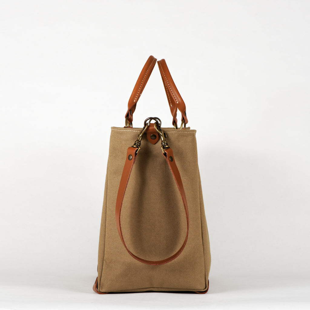 A beige cotton tote bag with brown leather handles and a removable brown leather strap, featuring a zippered closure and an interior pocket, set against a white background, side view.
