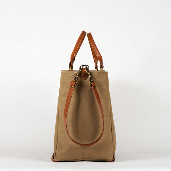 A beige cotton tote bag with brown leather handles and a removable brown leather strap, featuring a zippered closure and an interior pocket, set against a white background, side view.