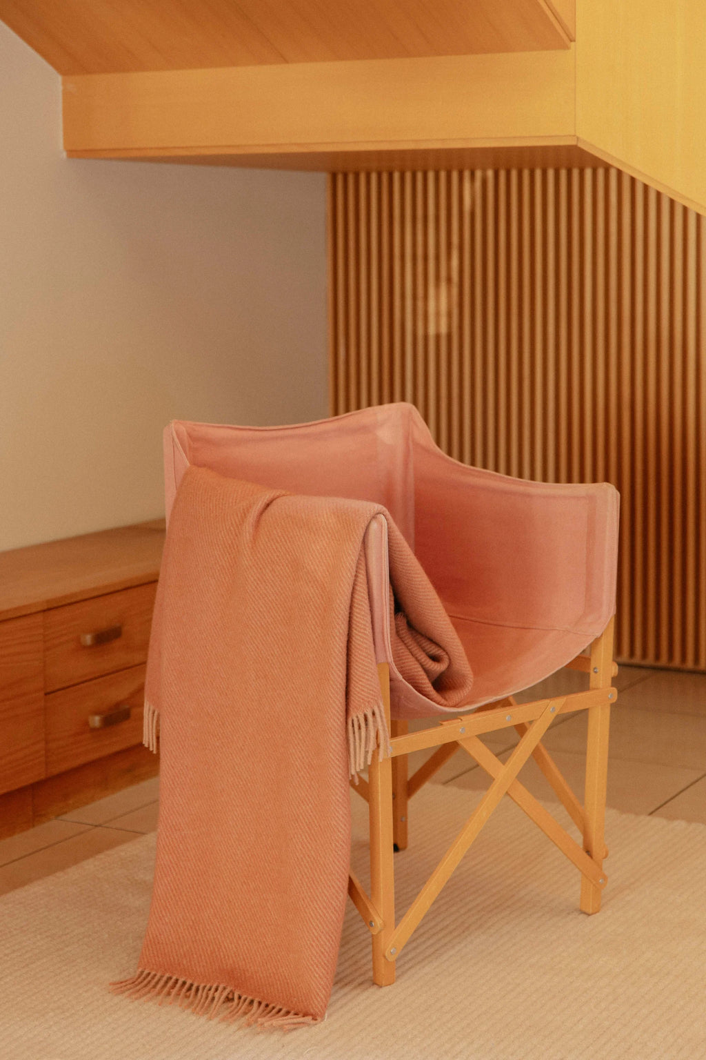 Powder cinnamon blanket draped over a wooden chair in a room with wooden furniture and a slatted wall.