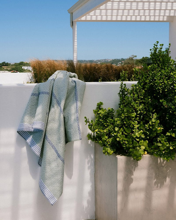 A bath towel with a geometric pattern hanging over a white wall with a lush green bush and a structure in the background.