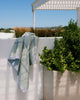 A bath towel with a geometric pattern hanging over a white wall with a lush green bush and a structure in the background.