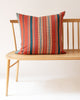 A red, blue, orange and white striped pillow displayed on a wooden bench with a white background.
