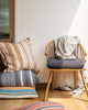 A variety of pillows with different patterns and colors displayed on a wooden chair and the floor, with natural lighting from a window in the background.