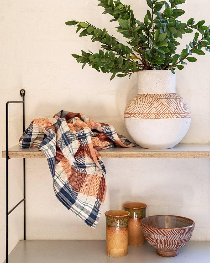 A checkered kitchen cloth in shades of orange and white is draped over a shelf, with a hanging loop visible. The shelf also holds a white vase with greenery and various kitchen items.