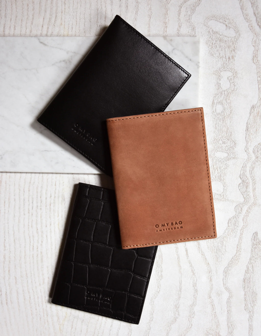 A collection of passport holders in black, brown, and dark brown colors, displayed on a light background. The brown holder has 'O My Bag' embossed on it.