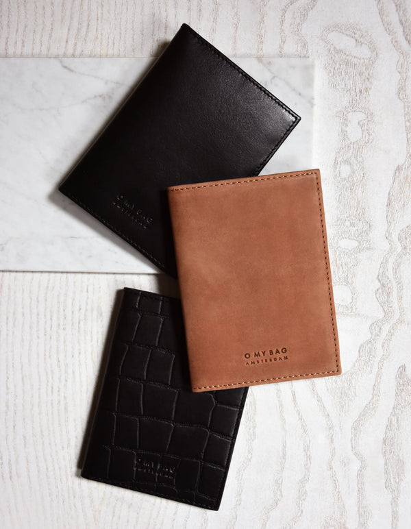 A collection of passport holders in black, brown, and dark brown colors, displayed on a light background. The brown holder has 'O My Bag' embossed on it.