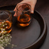 Hand holding a glass of amber liquid on a dark wooden tray with another glass and small plant.