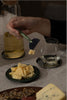 Person using a cheese knife to cut cheese on a plate with a glass of beer and another glass in the background.
