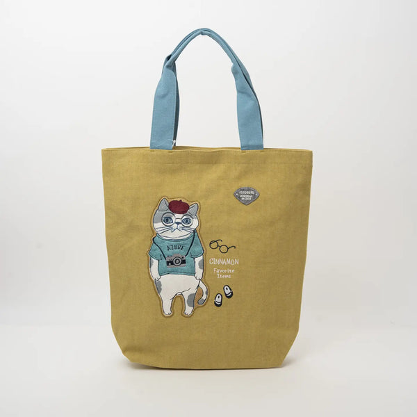 A mustard yellow canvas tote bag with light blue handles, featuring a cartoon illustration of a cat wearing a blue sweater and red beret, with the text 'CHINAMON' 