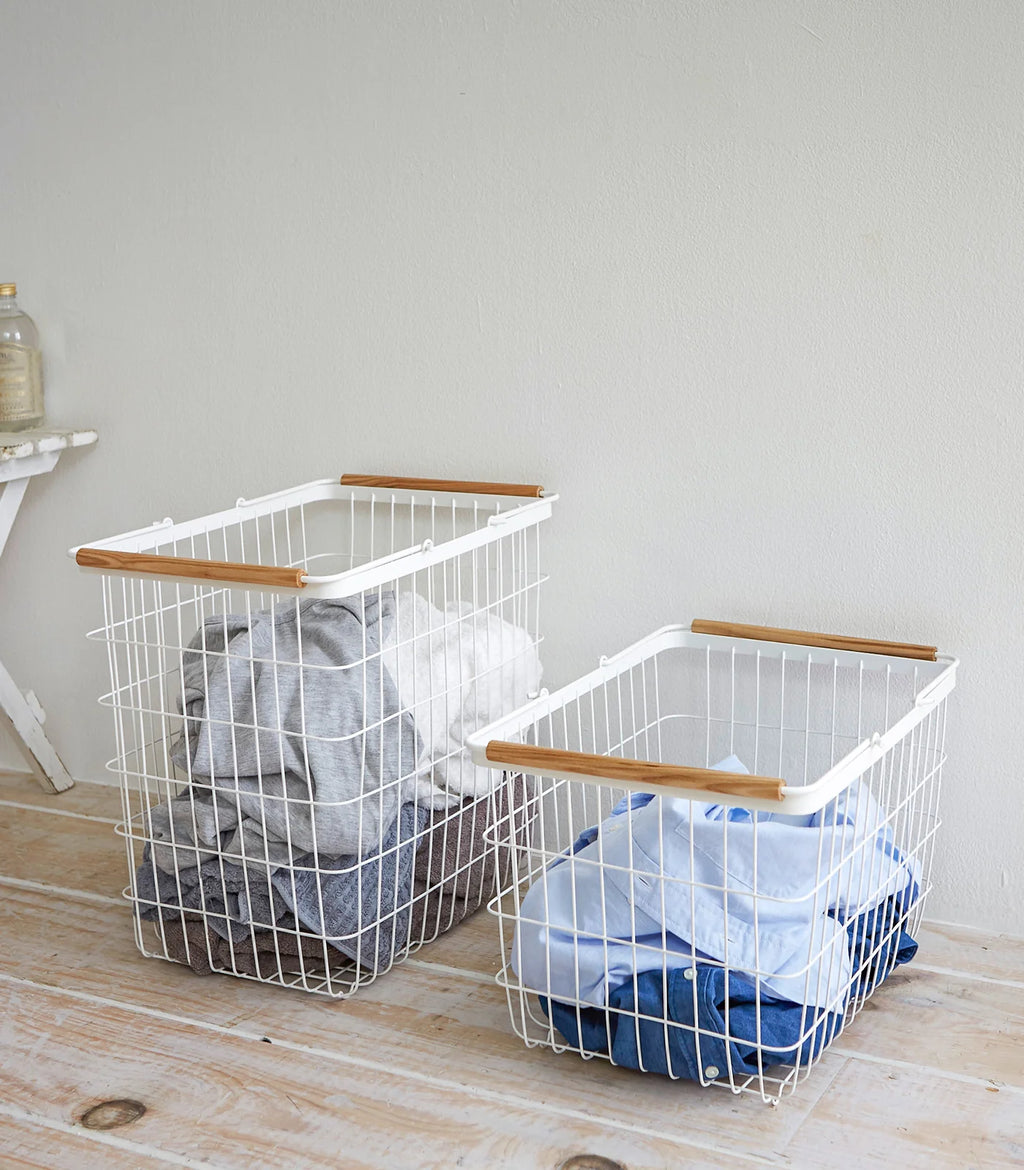 Two white wire baskets with wooden handles on a wooden floor against a light gray wall.