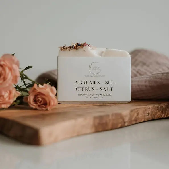 A bar of natural soap with placed on a wooden surface with decorative elements around it. Orange/blush roses in the background