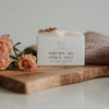A bar of natural soap with placed on a wooden surface with decorative elements around it. Orange/blush roses in the background
