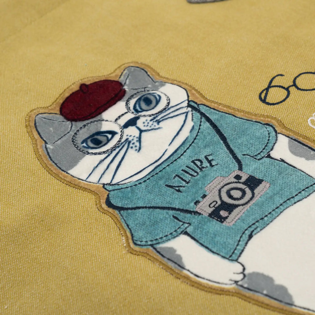 A close-up of a mustard yellow fabric featuring a detailed embroidered patch of a white cat wearing a teal shirt, a dark red beret, and a camera.
