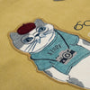 A close-up of a mustard yellow fabric featuring a detailed embroidered patch of a white cat wearing a teal shirt, a dark red beret, and a camera.
