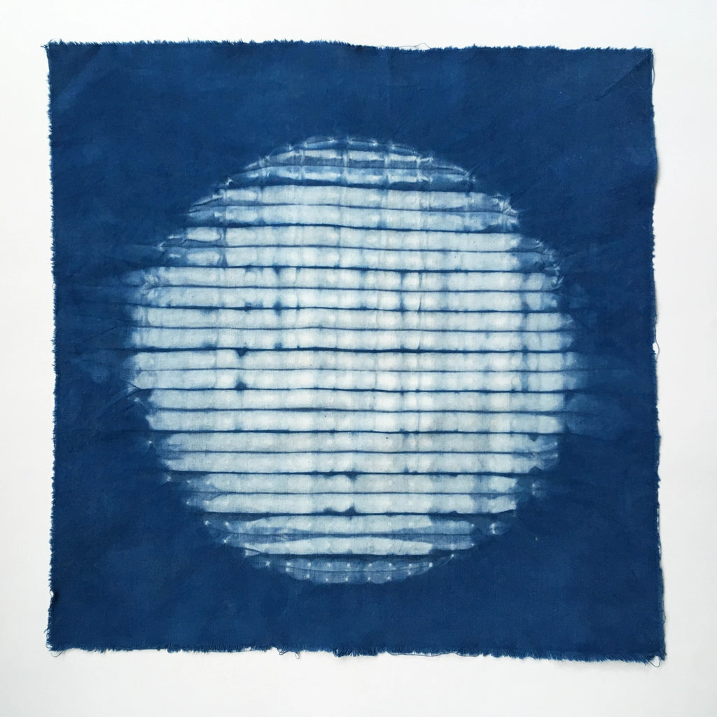 A square indigo blue fabric square with a white tie-dye shibori circle in the center, displayed flat against a white background.