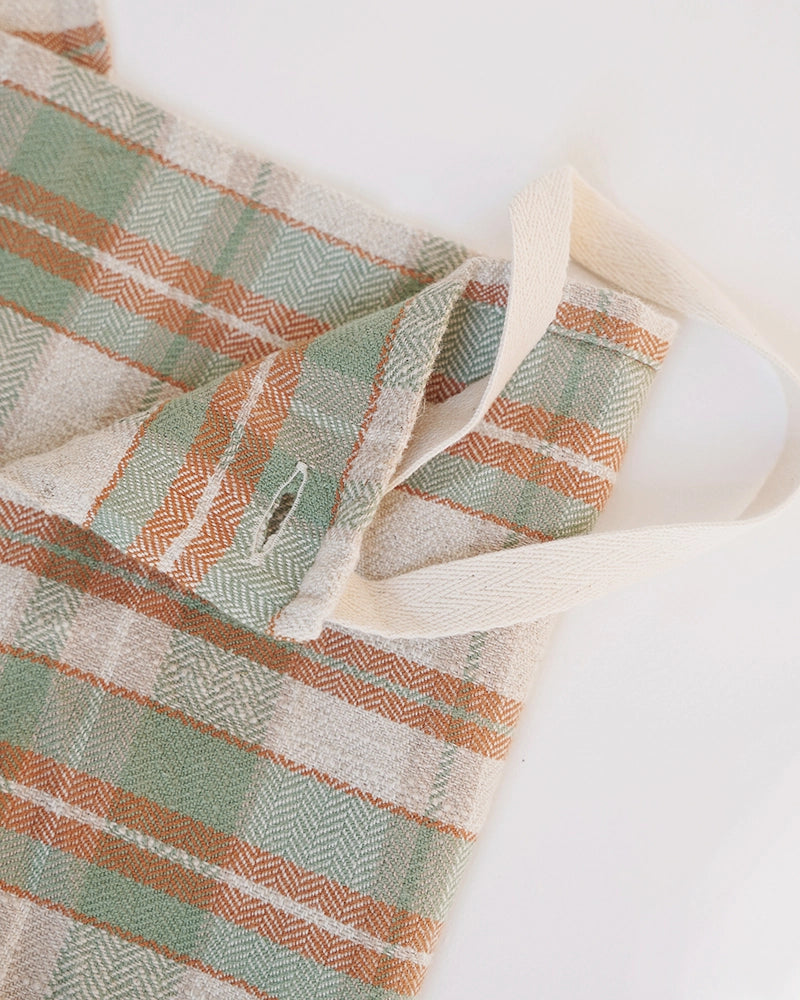 A close-up of a kitchen cloth with a muted plaid pattern in sage green, terracotta orange, and cream, featuring a cream canvas strap.