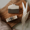 A bar of natural soap with 'ORANGE + PATCHOULI' labeled on the front, placed on a wooden surface with decorative elements around it.