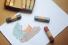 A set of pastel chalk sticks with beige and wood-toned bands, shown with a partially filled box and a white sheet of paper featuring painted color swatches in mint, peach, and beige on a brown surface.