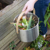 Person holding a metal bucket filled with kitchen scraps next to a wooden fence and plants.