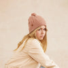 Woman wearing a blush pink beanie and beige jacket against a beige background